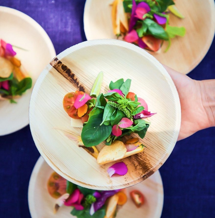 Palm leaf plates UK — round areca palm leaf plates with floral salad service (edible flowers, fresh herbs, baby vegetables, heirloom tomato), event catering presentation for UK wholesale distributors, supermarket own-label and foodservice procurement
