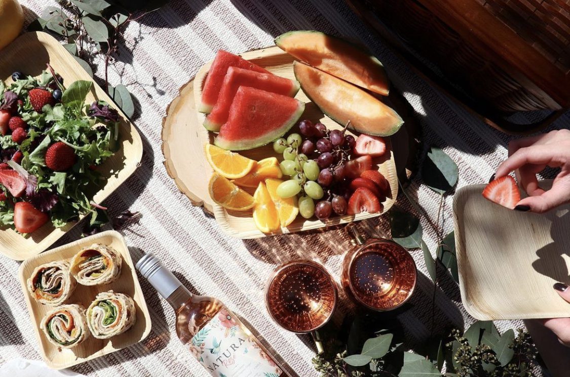 Areca palm leaf plates and bowls in lifestyle picnic service — salad, fresh fruit platter, and wrap rolls plated for outdoor dining, illustrating Ecodyne wholesale tableware in real HoReCa use for German distributors and event caterers